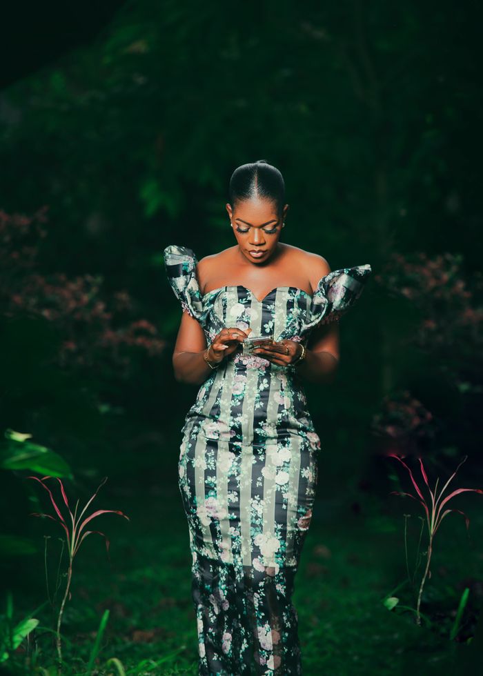 Beautiful African woman in an elegant floral dress standing in a lush green outdoor setting, radiating grace.
