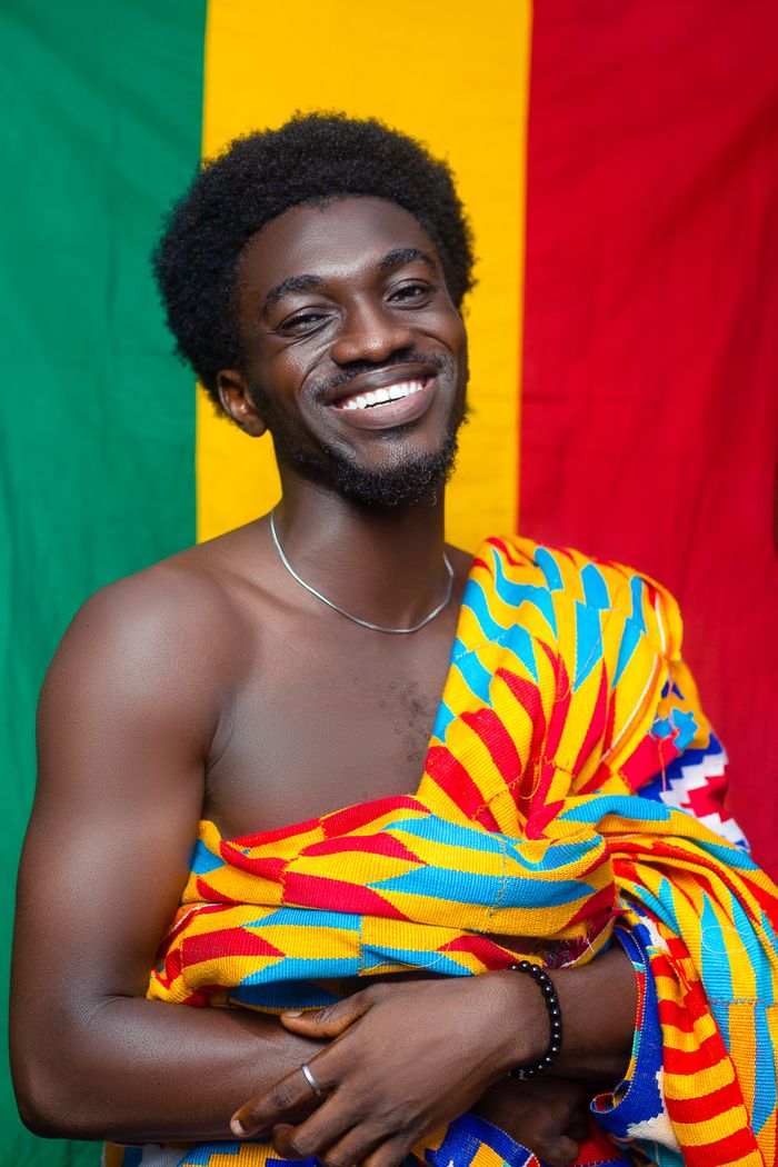 Man smiling wearing vibrant Kente cloth against colorful background.