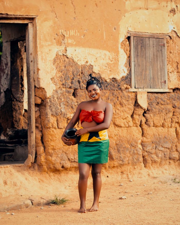 Smiling woman in Ghanaian dress holds pot outdoors, showcasing cultural heritage.