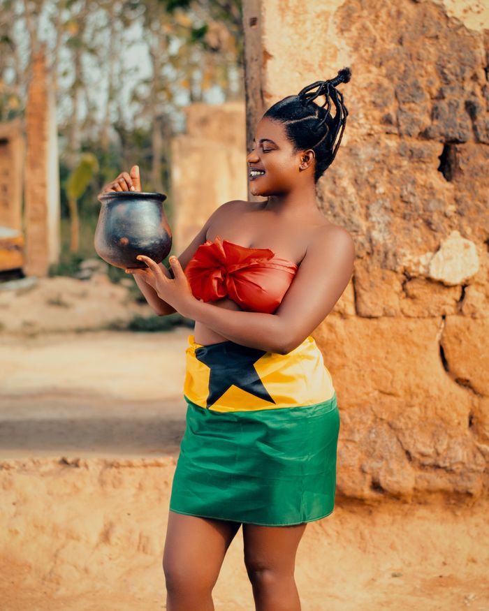 Woman in vibrant traditional dress holding pot, celebrating Ghana's culture outdoors.
